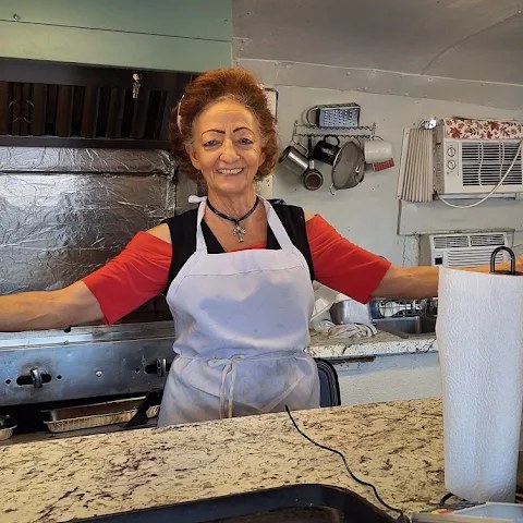 Maria smiling behind the service window of the Beachside Gyros truck, arms spread wide in her apron